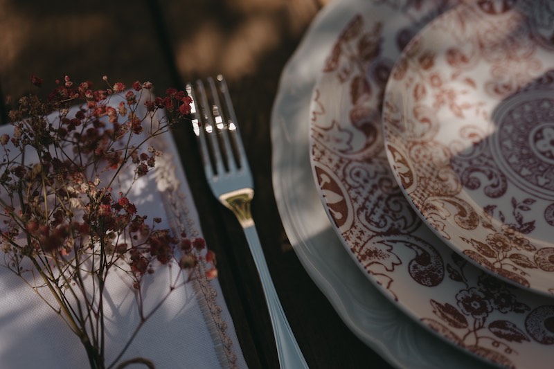 In questa foto il dettaglio di una mise en place di matrimonio con tavolo in legno nudo, piatti con decorazione di maioliche siciliane di colore rosso, tovagliolo in lino e fiori di campo secchi di colore rosso