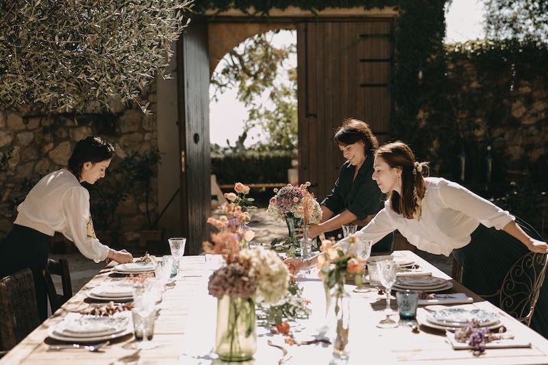 In questa foto la Wedding Planner Tania Costantino insieme a due collaboratrici sistema gli ultimi dettagli di una mise en place per matrimonio allestito nel cortile di un casale. Sullo sfondo si intravede un ulivo e il portone in legno del casale circondato da piante rampicanti. Il tavolo è decorato con un runner bianco, vasi, pampas, fiori di campo, sedie di diversa tipologia