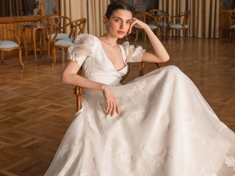 In questa foto una modella seduta su una poltrona in una sala in stile Liberty indossa un abito da sposa in tulle ricamato della collezione More 2023