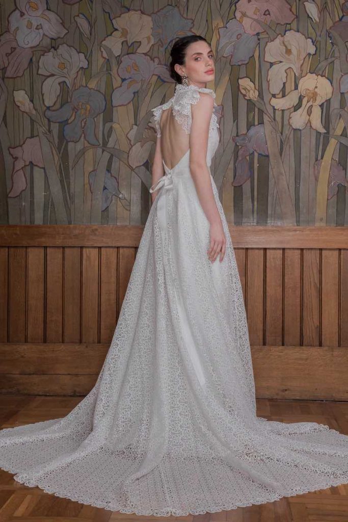 In questa foto una modella ritratta di spalle indossa un abito da sposa dalla linea svasata in macramè di cotone, scollo ad oblò sulla schiena e volant sulle maniche corte. Un piccolo fiocco stringe la vita sul retro