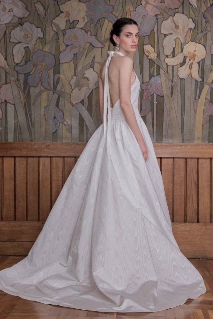 In questa foto una modella indossa un abito da sposa in seta moire della collezione More 2023. L'abito presenta una scollatura all'americana legata al collo con un nastro sottile e con schiena scoperta 