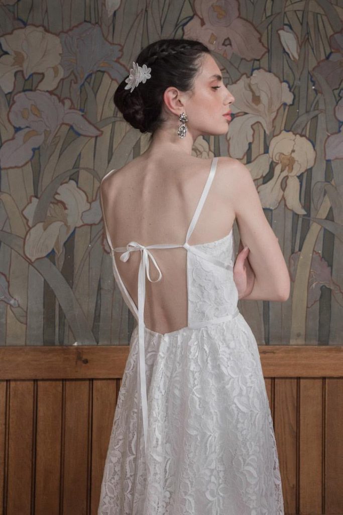 In questa foto una modella inquadrata di spalle indossa un abito da sposa scivolato More 2023 in pizzo Chantilly con scollatura geometrica sulla schiena creata da sottili nastri di seta 