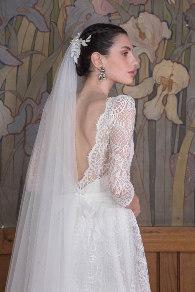 In questa foto una modella ritratta di spalle e a mezzo busto indossa un abito da sposa More in pizzo chantilly con maniche a tre quarti e scollatura sulla schiena. La modella indossa anche un velo in tulle con fermaglio floreale 