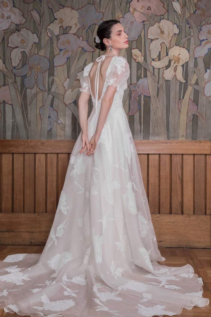 In questa foto una modella inquadrata di spalle indossa un abito da sposa More 2023 in tulle ricamato con motivi floreali e scollatura ad oblò sulla schiena chiusa da un piccolo fiocco