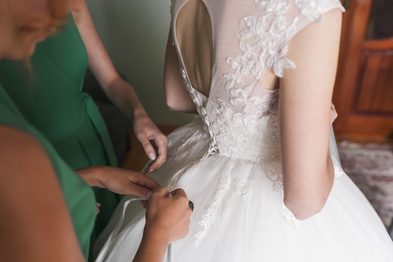 In questa foto il dettaglio di una sposa di spalle mentre le viene sistemato l'abito sul retro