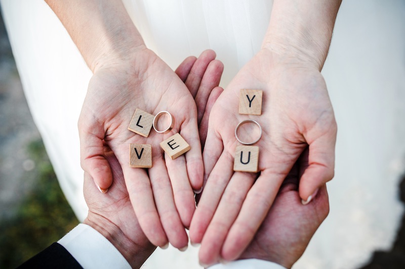 In questa foto due mani aperte l'una sull'altra con la scritta "Love" composta da tasselli di legno di forma quadrata dove le O sono sostituite da due fedi nuziali