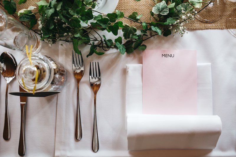 In questa foto un tavolo per matrimonio fotografato dall'alto e allestito con edera, runner di juta, tovagliato bianco e un menù in carta di colore rosa