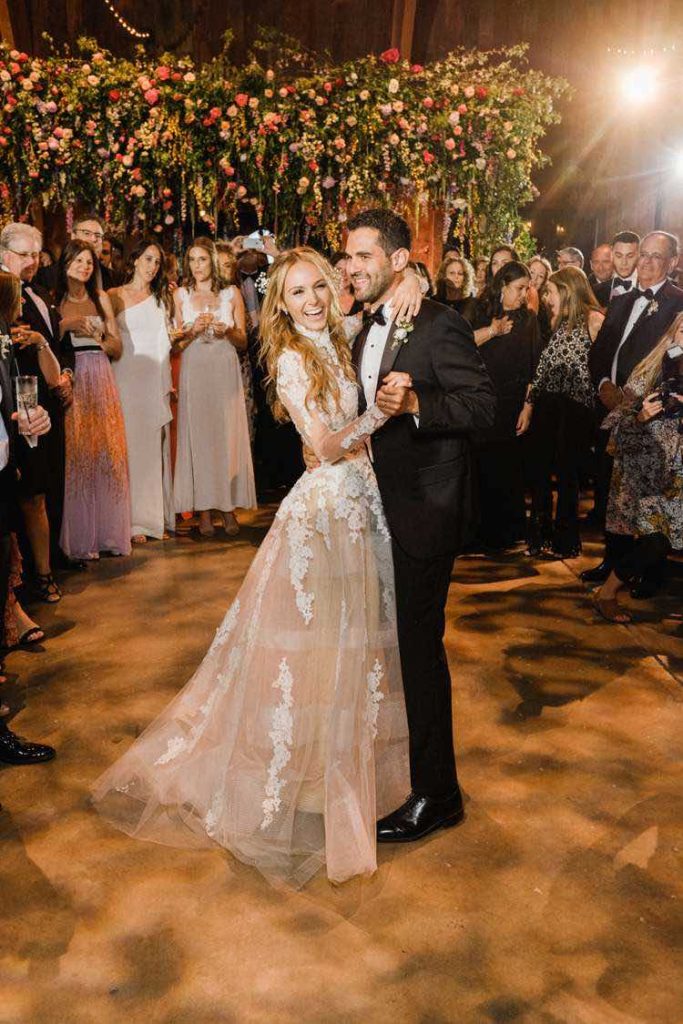 In questa foto una sposa e uno sposo sorridono al fotografo durante il loro primo ballo sposi. Lei indossa un abito in tulle con ricami floreali ramage, maniche lunghe e collo alto. Lo sposo indossa uno smoking. Attorno a loro sorridono e battone le mani tutti gli ospiti del matrimonio