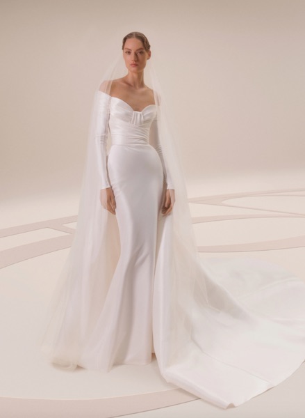 In questa foto una modella indossa un abito da sposa sirena in mikado di seta off white, con scollatura bateau e maniche lunghe. L'abito ha un leggero drappeggio sul corpetto
