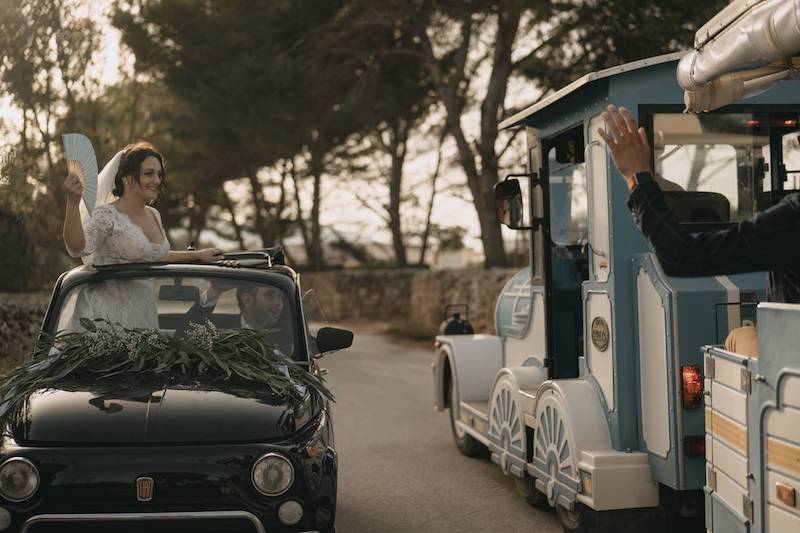 In questa foto la sposa Pinuccia in piedi nella Cinquecento guidata da Andrea mentre con un ventaglio colore azzurro polvere saluta i suoi ospiti a bordo di un trenino
