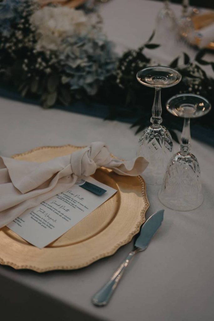 In questa foto il dettaglio di una mise en place con sottopiatto di colore oro, tovagliolo di lino, menù, bicchieri di vetro