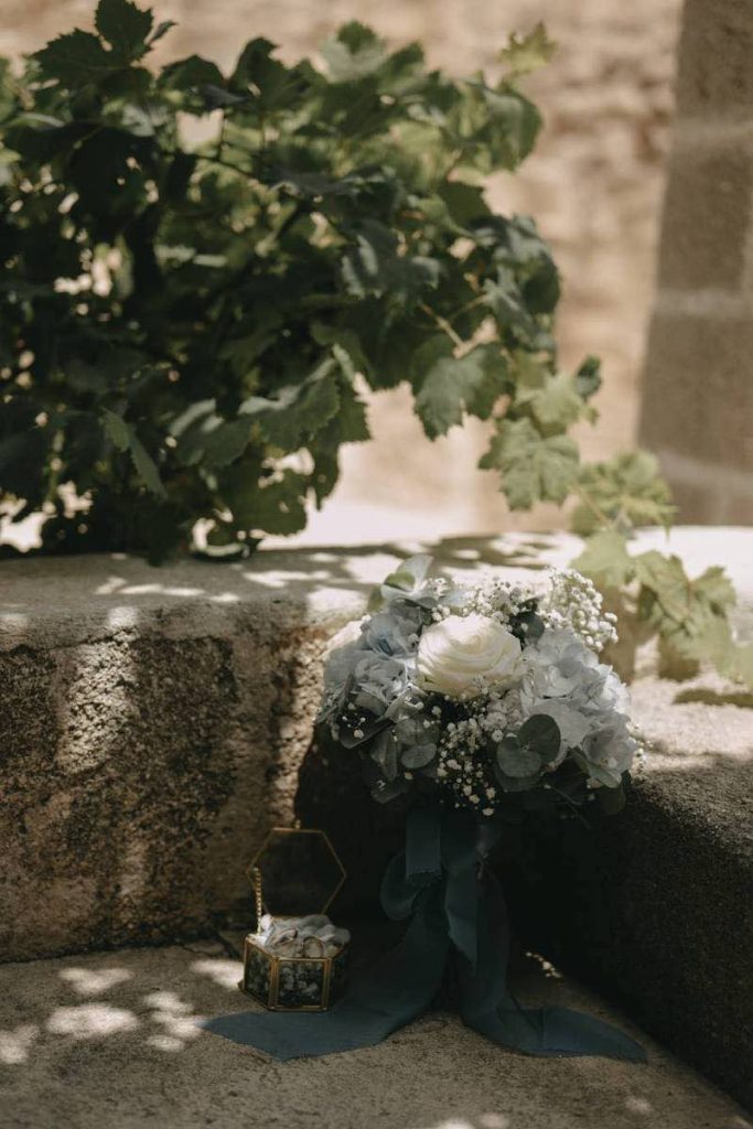 In questa foto il dettaglio del bouquet con rose di colore bianco, ortensie di colore azzurro e foglie di eucalipto chiusi da nastri di colore azzurro polvere. Accanto al bouquet è posizionato il portafedi a forma ottagonale 