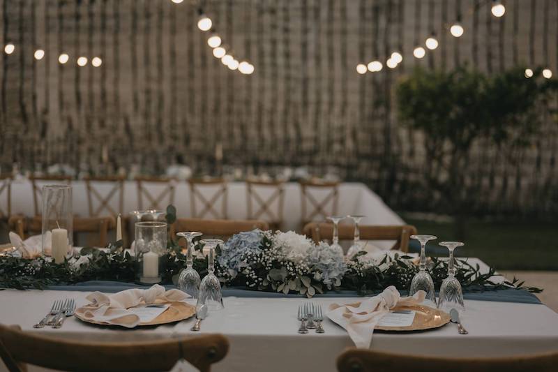 In questa foto il dettaglio di una mise en place per matrimoniocomposta da una tovaglia di colore bianco, sottopiatti di colore oro, bicchieri di vetro, e un runner di lino di colore azzurro decorato con candele e rose bianche e 