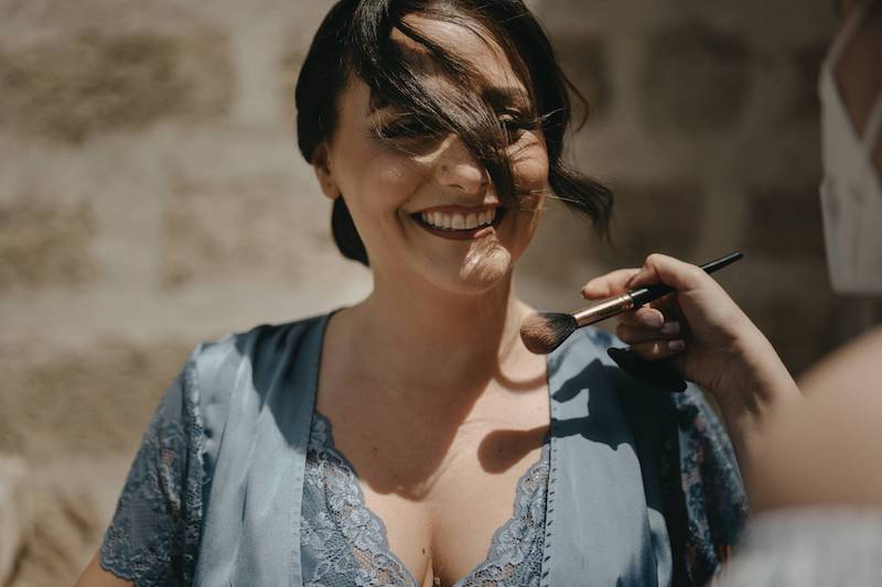 In questa foto un primo piano della sposa Pinuccia in lingeria colore azzurro polvere con i capelli raccolti e un ciuffo che le passa sul viso mentre viene truccata all'aperto