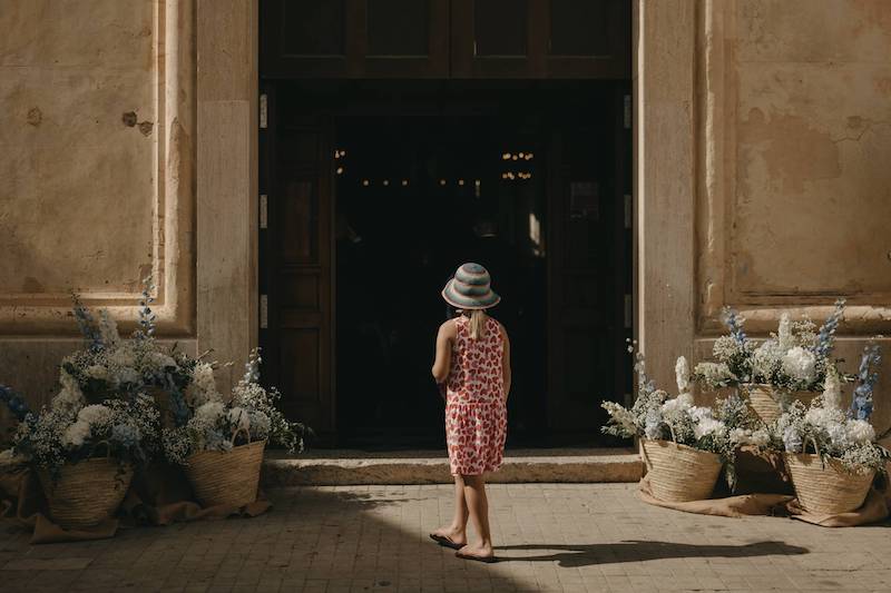 In questa foto una bambina di spalle mentre guarda l'ingresso della chiesa di un matrimonio. L'ingresso è decorato con coffe di paglia che contengono ortensie, rose e delphinium di colore azzurro