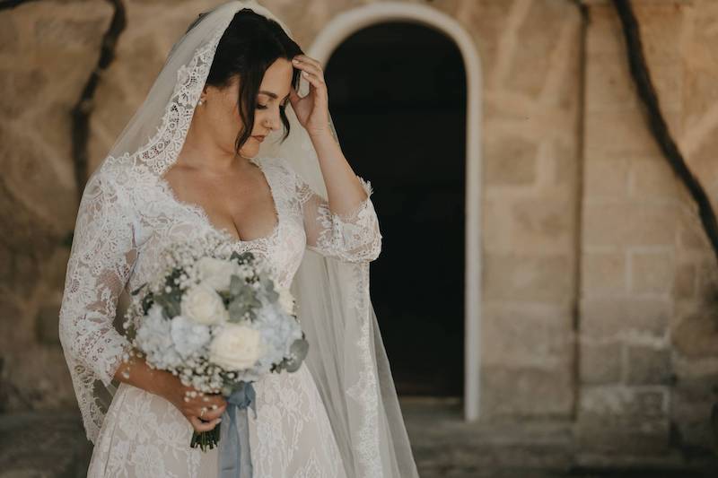 In questa foto la sposa Pinuccia con un vestito da sposa interamente in pizzo velo a mantiglia abbinato. Tiene tra le mani un bouquet con rose di colore bianco ortensie di colore azzurro fermati da nastri abbinati