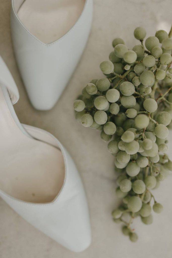 In questa foto il dettaglio di un paio di scarpe da sposa a punta e un grappolo di uva bianca