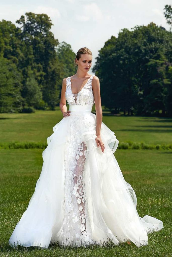 In questa foto una modella indossa un abito da sposa Stefano Blandaleone 2023 con spacco con fiori 3D e sovragonna a balze. Gli stessi fiori sono presenti sul bustino a v fermato da una cintura di seta