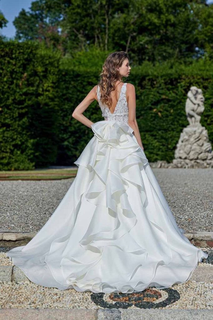 In questa foto una modella inquadrata di spalle indossa un abito da sposa Stefano Blandaleone 2023 con bustino con scollo a V sulla schiena, fiori 3D e strascico con multi balze