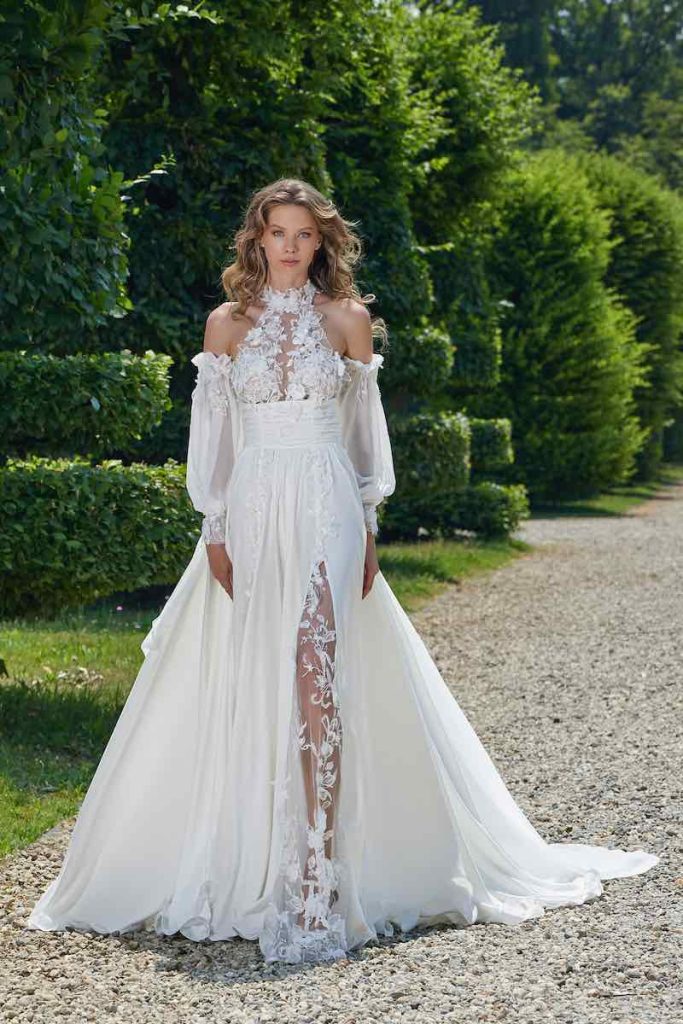 In questa foto una modella indossa una abito da sposa scivolato Stefano Blandaleone con gonna in georgette con spacco in tulle ricamato. L'abito presenta una scollatura all'americana con fiori 3D, una fascia in seta drappeggiata e maniche a sbuffo in organza di seta