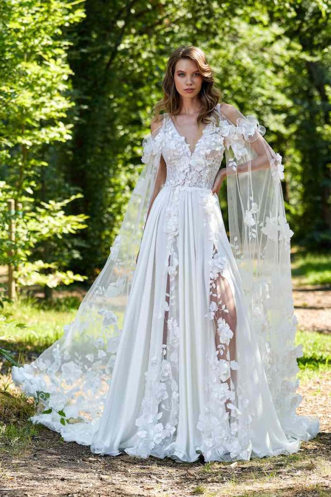 In questa foto una modella indossa un abito da sposa Stefano Blandaleone 2023 dalla linea scivolata con fiori 3D e ali di tulle ricamate con fiori 3D. La gonna in georgette presenta due spacchi frontali