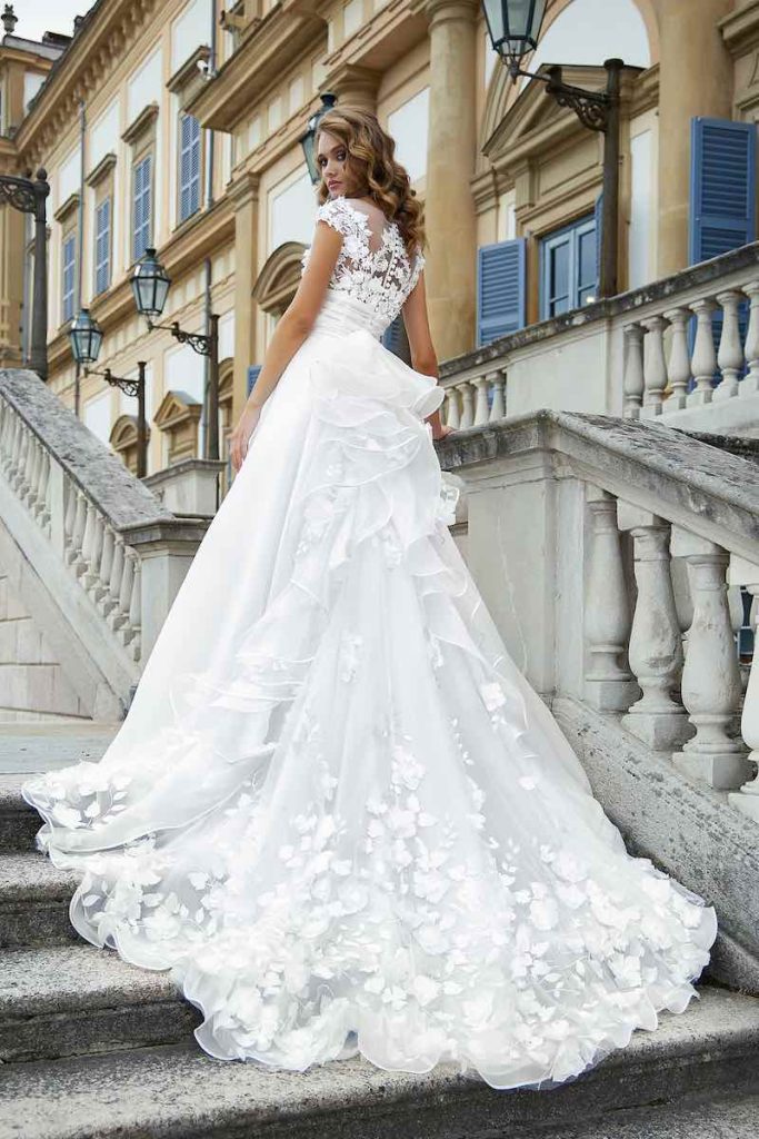 In questa foto una modella inquadrata di spalle sulle scale di un palazzo nobiliare indossa un abito da sposa Stefano Blandaleone 2023 con strascico floreale e rouches che partono dalla vita 