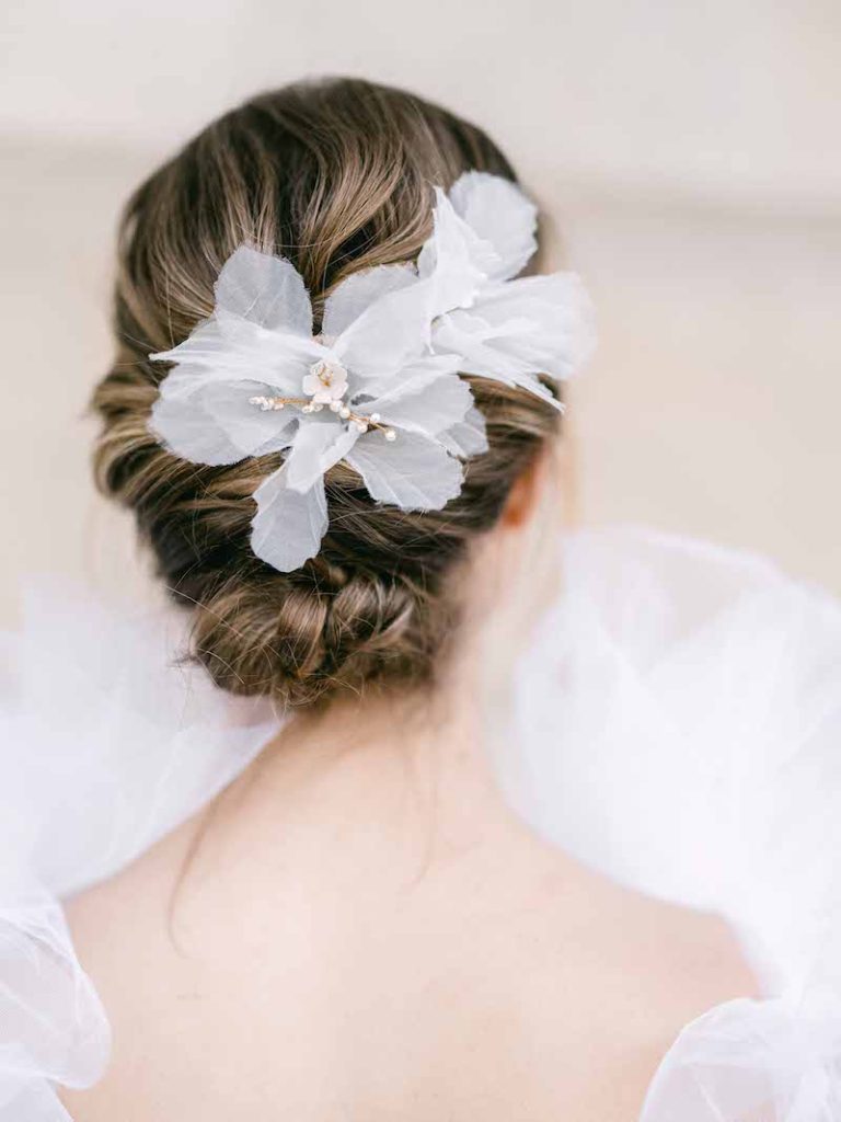 In questa foto una sposa di spalle mostra una raccolto semplice decorato da maxi fiori di tessuto trasparente di colore bianco decorati con piccoli fiorellini smaltati