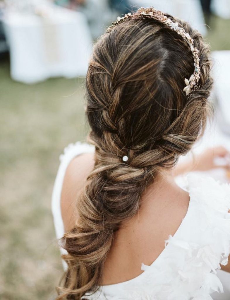 In questa foto una sposa di spalle mostra una delle acconciature sposa 2023 più di tendenza: una treccia fermata da una tiara gioiello