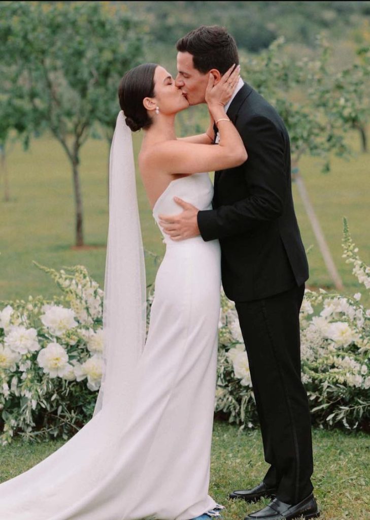 In questa foto due sposi di profilo si baciano. La sposa tiene tra le mani il viso dello sposo e indossa un abito a sirena liscio, un velo a fazzoletto fermato su uno chignon basso