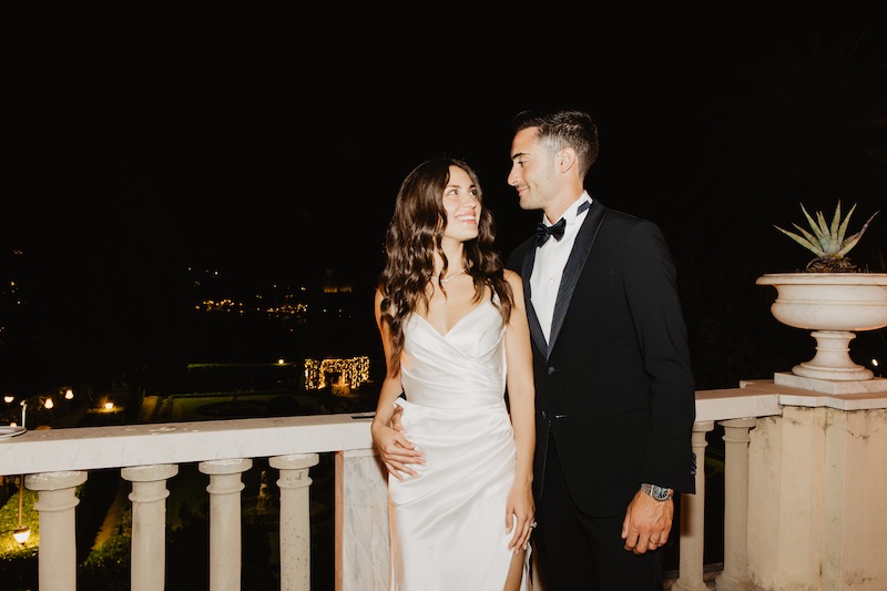 In questa foto una sposa abbracciata allo sposo davanti alla balaustra in marmo di una terrazza di sera mostra il suo cambio da abito in satin drappeggiato e il cambio di acconciatura con onde morbide effetto bagnato