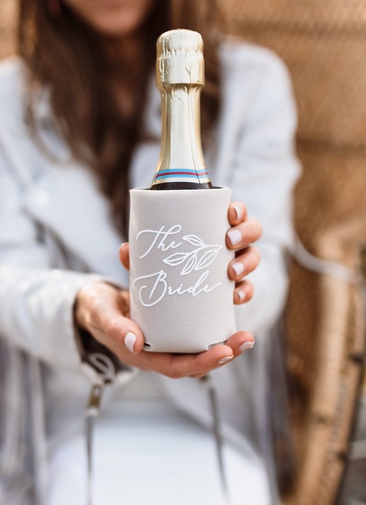 In questa foto le mani di una ragazza che tiene in mano una mini bottiglia di champagne dentro ad un Wedding Koozie personalizzato con una scritta di colore bianco "The Bride"