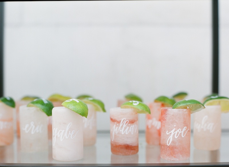 In questa foto shot con cocktail di colore rosa pesca usati come escort card. I bicchieri riportano i nomi degli ospiti e sulle fettine di limone il numero del tavolo