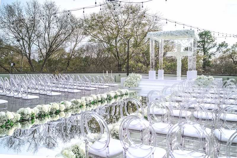 In questa foto l'allestimento di un matrimonio civile composto da sedie in plexiglass trasparente, un corridoio specchiato decorato sui bordi da composizioni di fiori di forma rotonda e di colore bianco. Alla fine del corridoio è posizionato un gazebo decorato con fiori di colore bianco