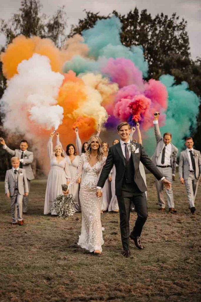 In questa foto due sposi camminano su un prato tenendosi mano nella mano mentre damigelle e testimoni agitano in aria le bombe di fumo colorato