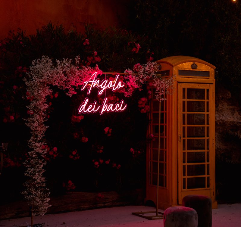 In questa foto una delle idee matrimonio 2023: l'angolo dei baci con scritta al neon appesa ad una cornice decorata con gypsophila colorata e posizionata accanto ad una cabina del telefono vintage e di colore giallo