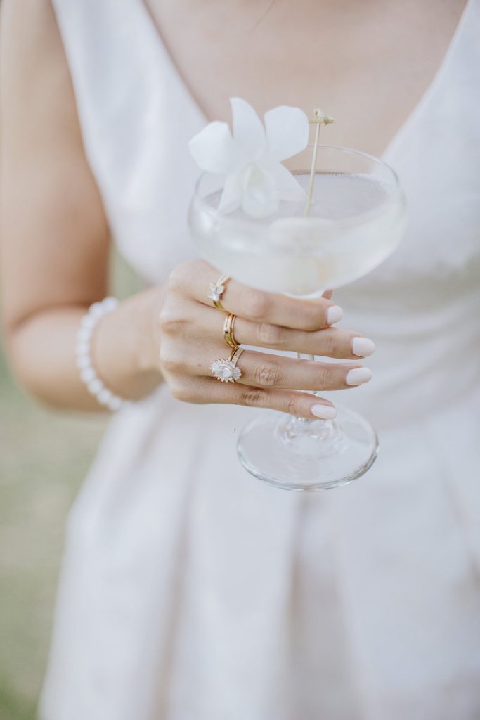 In questa foto la mano di una sposa con 3 anelli che tiene in mano un cocktail decorato con un fiore bianco