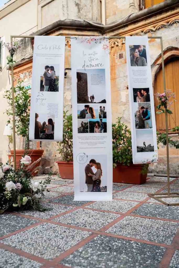 In questa foto una delle idee matrimonio 2023 più gettonate: il Photo Display composto da runner di carta rigida sul quale sono presenti foto degli sposi e il racconto della loro storia d'amore. I runner di carta sono appesi su una cornice rigida decorata con fiori di colore rosa e bianca