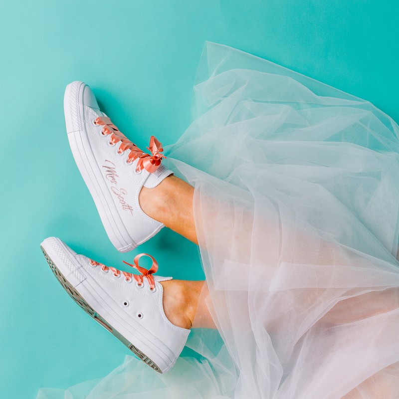 In questa foto il dettaglio di un paio di sneakers da sposa di colore bianco con lacci di seta di colore rosa pesca e il suo nome ricamato sul fianco
