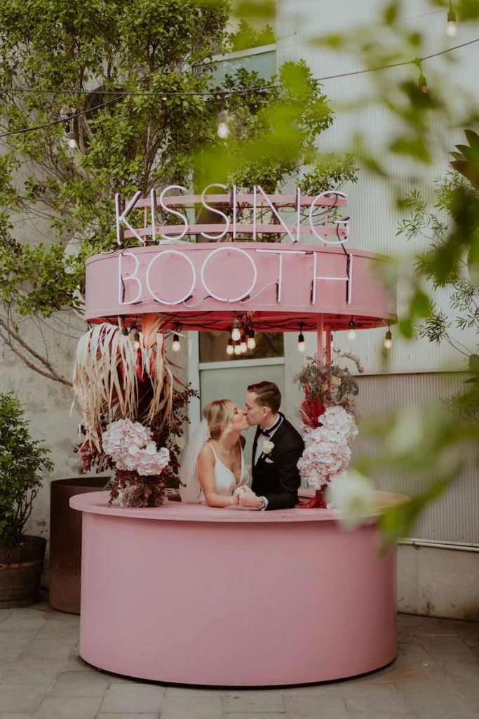 In questa foto due sposi si baciano dietro all'interno al Kissing Booth composto da un bancone rosa decorato con fiori di colore rosa e bianco e sotto una scritta al neon "Kissing Booth"