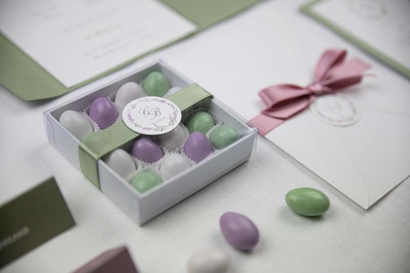 In questa foto una scatola di colore bianco con confetti di colore viola, bianco e verde. La scatola è chiusa da un nastro di colore verde decorato con un bollino con le iniziali degli sposi