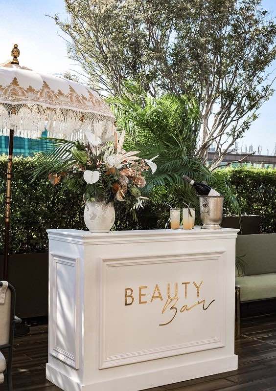 In questa foto il bancone di colore bianco della Beauty Bar di un matrimonio decorato con fiori e sul quale sono adagiati tre bicchieri con cocktail e un cestello con vino. Sulla facciata del bancone è riportata la scritta dorata "Beauty Bar