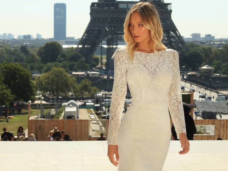 In questa foto una modella fotografata davanti alla Tour Eiffel inquadrata a mezzo busto indossa un abito da sposa in pizzo con maniche lunge di Iryna Kotapska