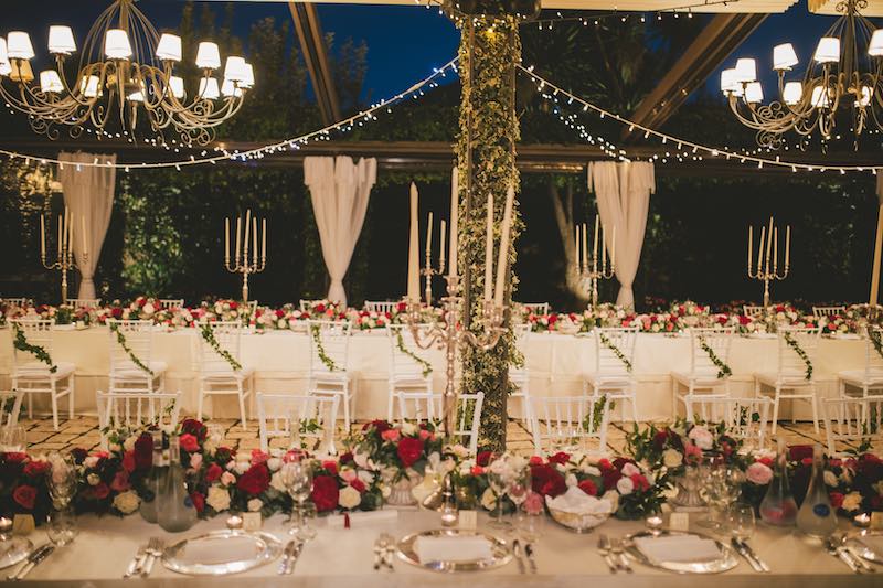 In questa foto un allestimento di matrimonio all'aperto realizzato con tavoli imperiali decorati con runner di fiori di colore rosso, rosa e bianco. Sopra i tavoli sono presenti grandi lampadari