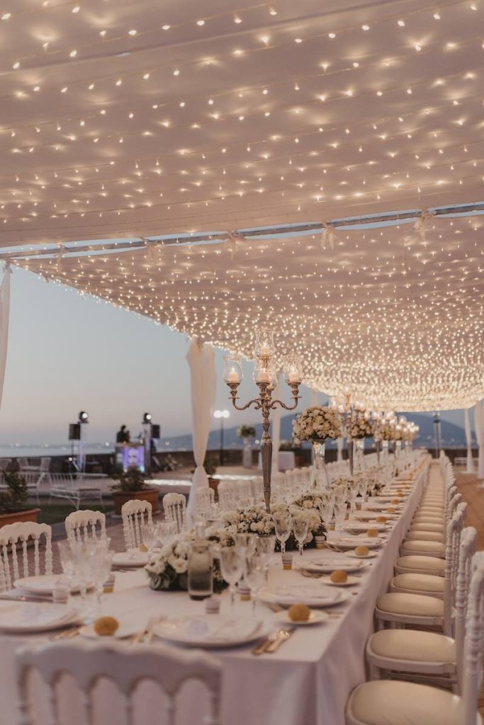 In questa foto l'allestimento di un matrimonio su una terrazza realizzato con tetto di luci sopra un lungo tavolo imperiale decorato in bianco