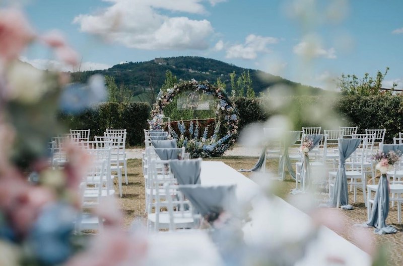 In questa foto un allestimento per la cerimonia di un matrimonio civile nei toni dell'azzurro polvere, del rosa e del bianco. Sulle sedie chiavarine sono adagiati drappi di colore azzurro polvere fermati da composizioni di fior. Sullo sfondo è presente un cerchio di fiori abbinati