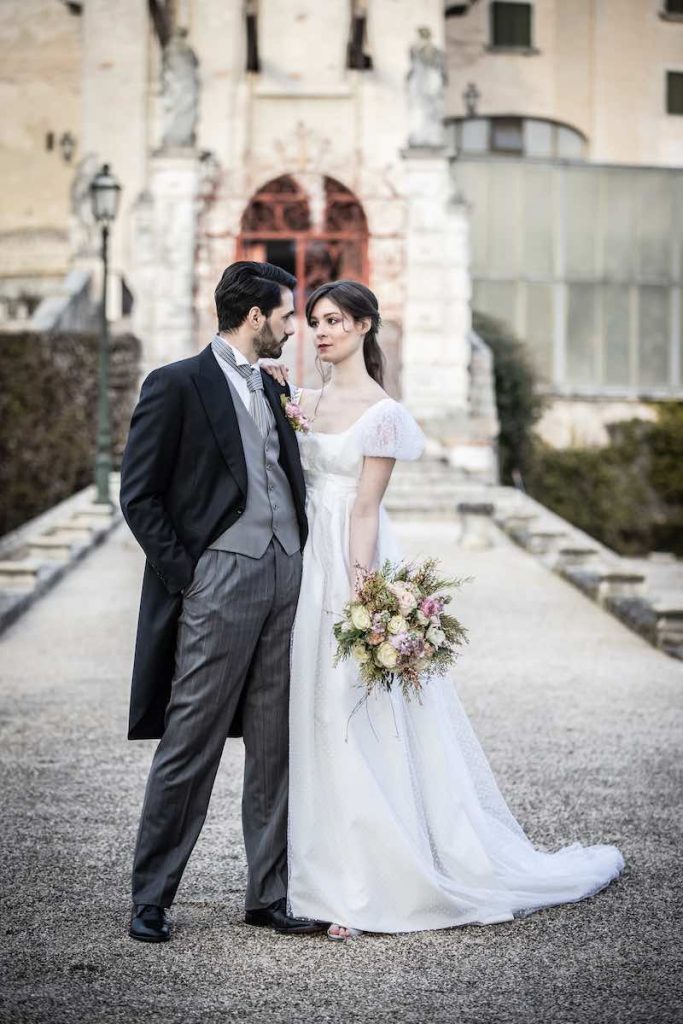 In questa foto due sposi si guardano abbracciati. La sposa indossa un abito stile impero con maniche balloon. Lo sposo indossa un tighte nel colore del grigio antracite e del grigio perla