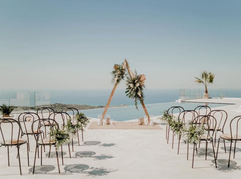 In questa foto l'allestimento di un matrimonio civile a picco sul mare realizzato con sedie di legno decorate con fiori bianchi e verdi. Sullo sfondo si intravede un backdrop di forma triangolare di legno decorato con composizioni di fiori e candele 