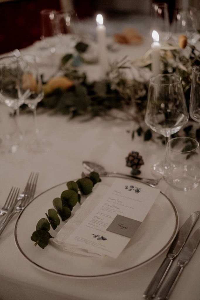 In questa foto il dettaglio di una mise en place nei toni del grigio, del bianco e del verde. In primo piano è mostrato un piatto decorato con un menù e un rametto di eucalipto