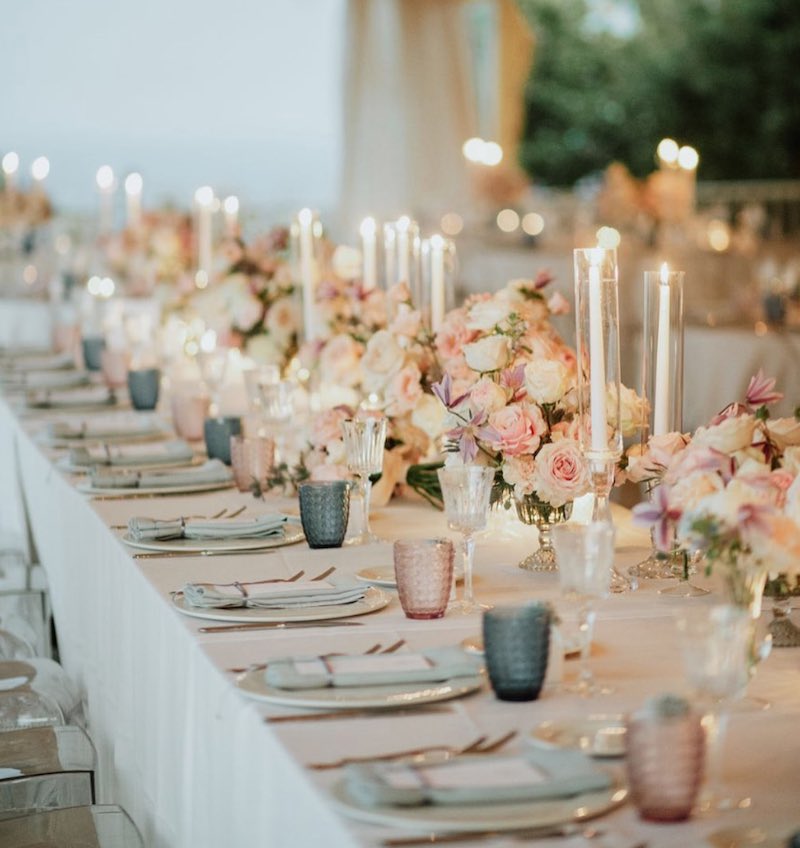In questa foto una mise en place per matrimonio nei toni pastello del rosa e dell'azzurro con composizioni di fiori in vasi lungo il tavolo imperiale e portacandele in vetro con candele bianche 