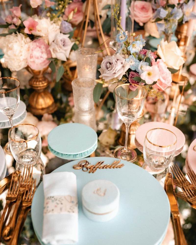 In questa foto il dettaglio di una mise en place per matrimonio in stile Bridgerton nei toni dell'oro, del rosa e del turchese. In primo piano è inquadrato un sottopiatto di colore turchese con scatolina cadeaux e segnaposto con scritta laser abbinate. Sullo sfondo sono presenti portavasi colore oro con fiori di colore turchese e rosa